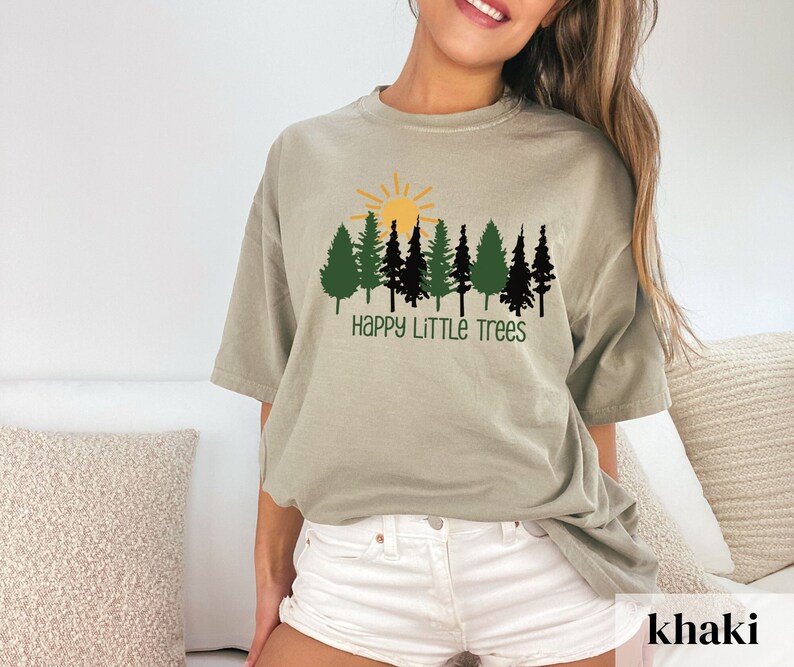 Woman wearing white shorts and a beige t shirt with a sunshine and trees and the text Happy Litte Trees in green.