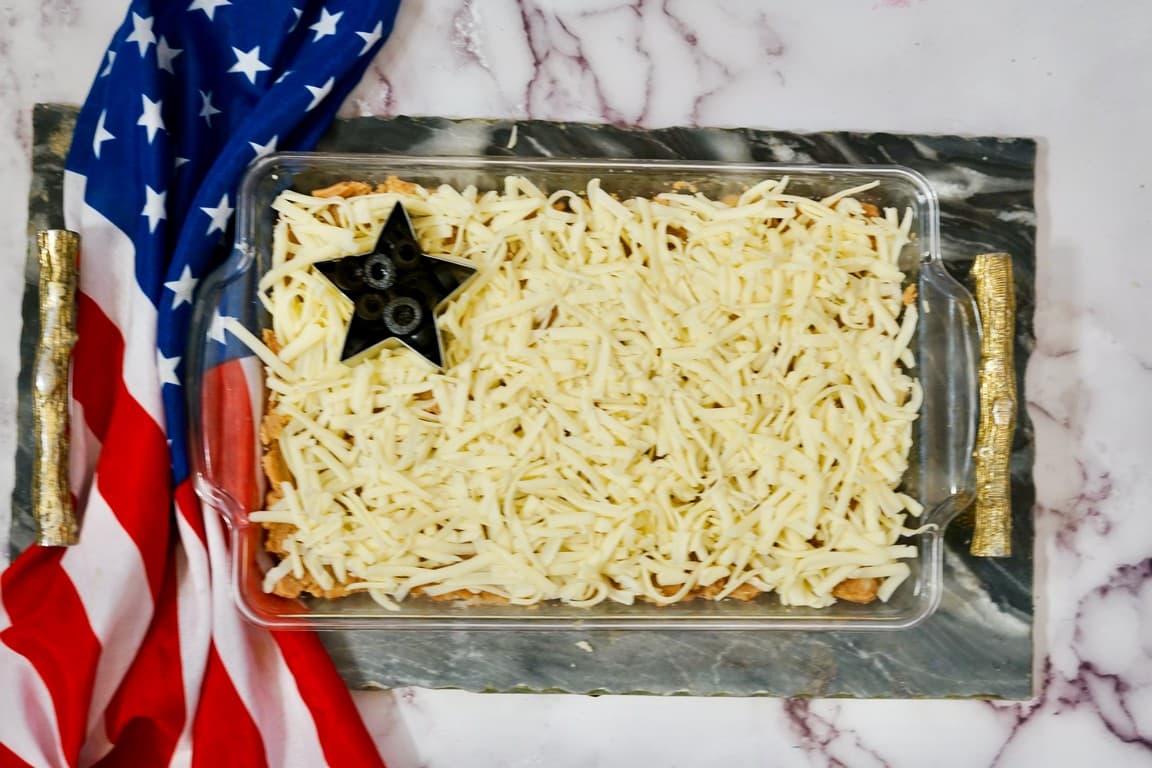 Glass pan with bean layer, cheese layer, and a star cookie cutter with black olives in it. 